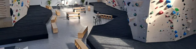 Salle d'escalade vide avec les différents murs de grimpes et leurs différents niveaux, indiqués par les couleurs noir, bleu, vert, jaune et rouge. Des matelas de réception sont disposés au bas des murs.